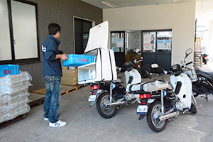 新聞配達バイク（右）の横で、食品専用バイクに森永商品を積み込む